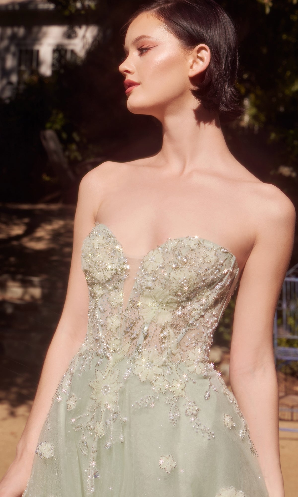 A woman stands outdoors in sunlight wearing the Sage Green Prom Ball Gown: Andrea & Leo A1418, a strapless corset dress adorned with floral embroidery and beading.